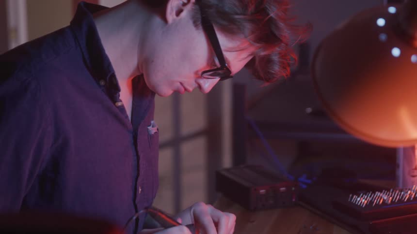 A young man wearing glasses concentrates while using a soldering iron on electronics at a workbench illuminated by colored light
