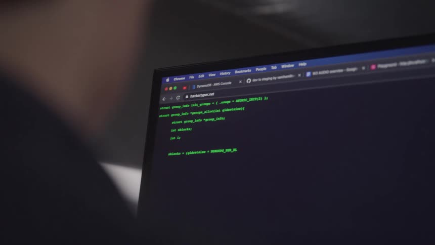 Viewed over someones shoulder lines of code automatically appear on a laptop screen displaying the website hackertypernet