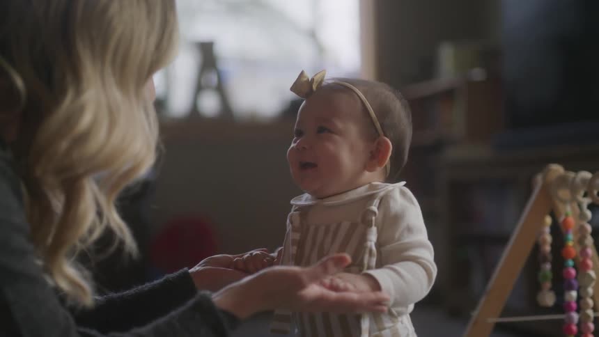 A woman with long blonde hair is holding the hands of a baby dressed in a striped outfit with a wooden baby gym featuring colorful beads in the background