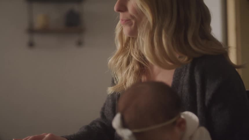 Sitting indoors at a table a blonde mother wearing a grey sweater holds her baby daughter who wears a white shirt and headband while using her fingers to scroll on a laptop placed in front of them