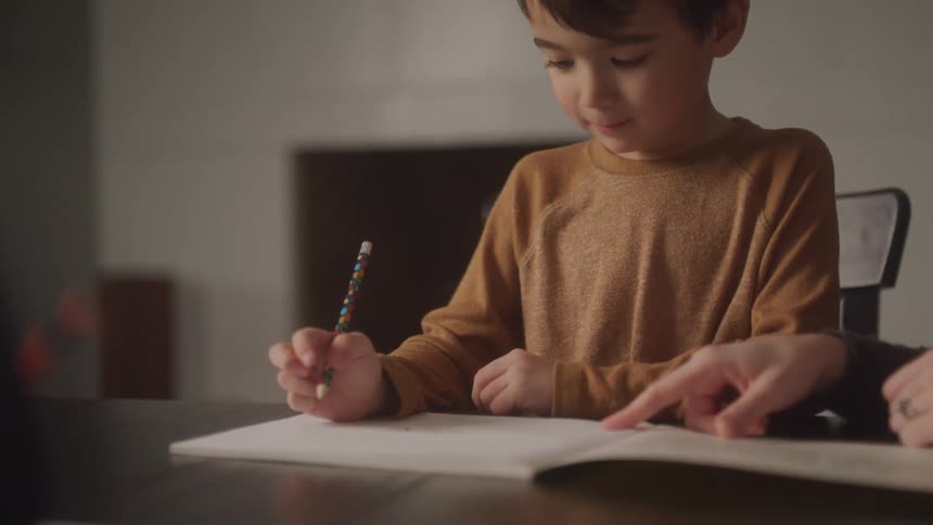 The child carefully works on homework with pencil in hand while his mother points to the page guiding him step by step at a simple table