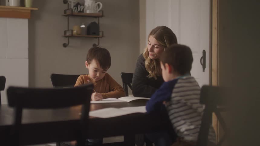 Gathered around a dark wood dining table indoors a mother attentively engages with her two young sons who are seated and working on their homework assignments