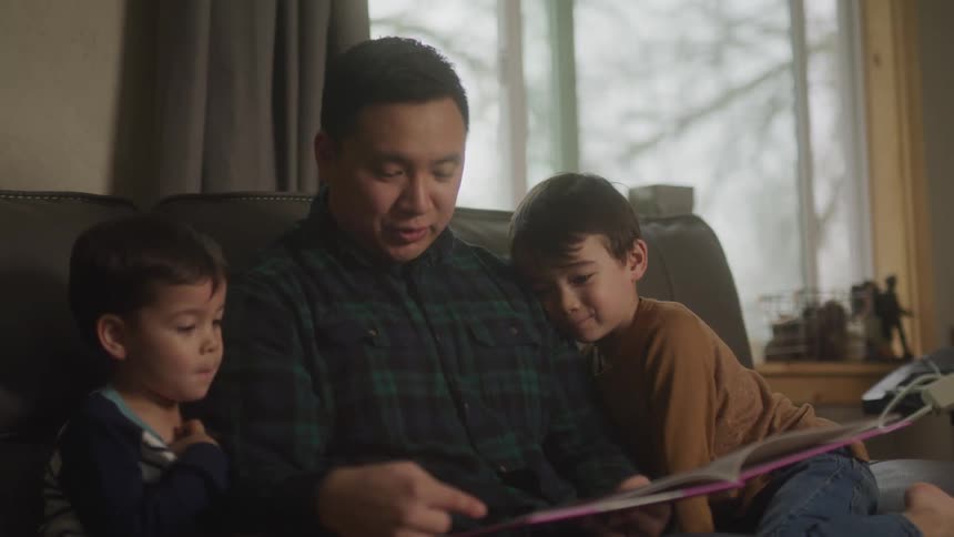 Father sitting on the couch in the living room reading a book to his two young children