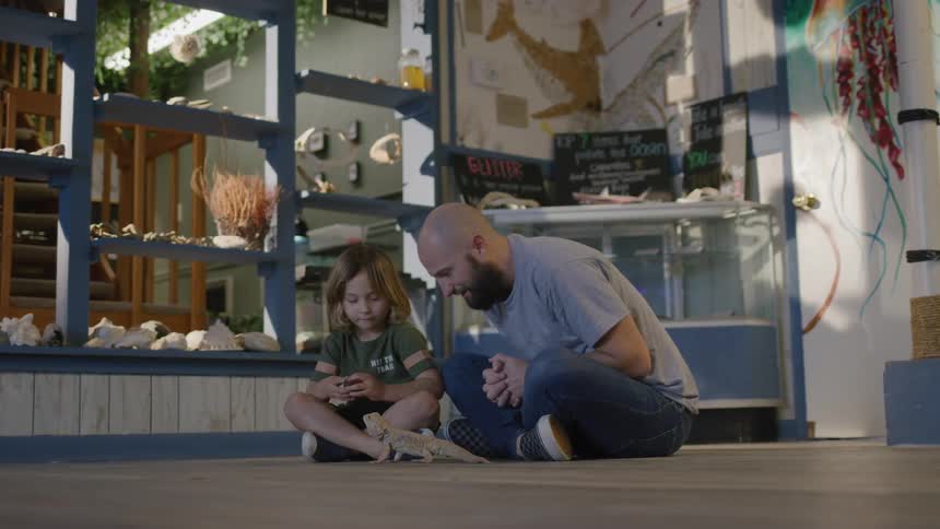 A father sits on the floor of a museum and teaches his son about reptiles