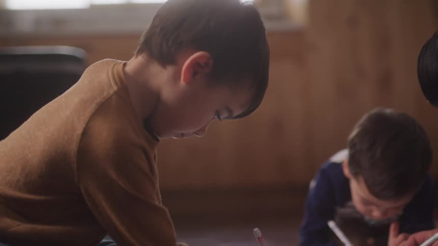 Two young boys with dark hair are sitting on the floor drawing or writing on papers in front of them