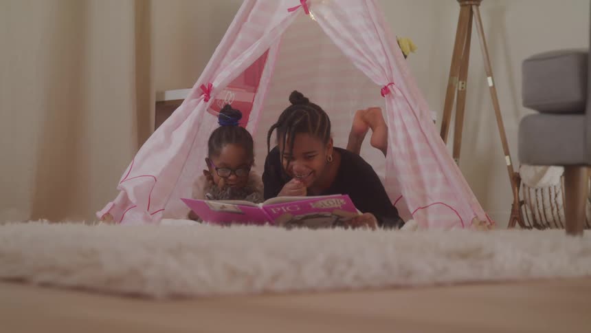 Two young girls are laughing while reading a book on the floor
