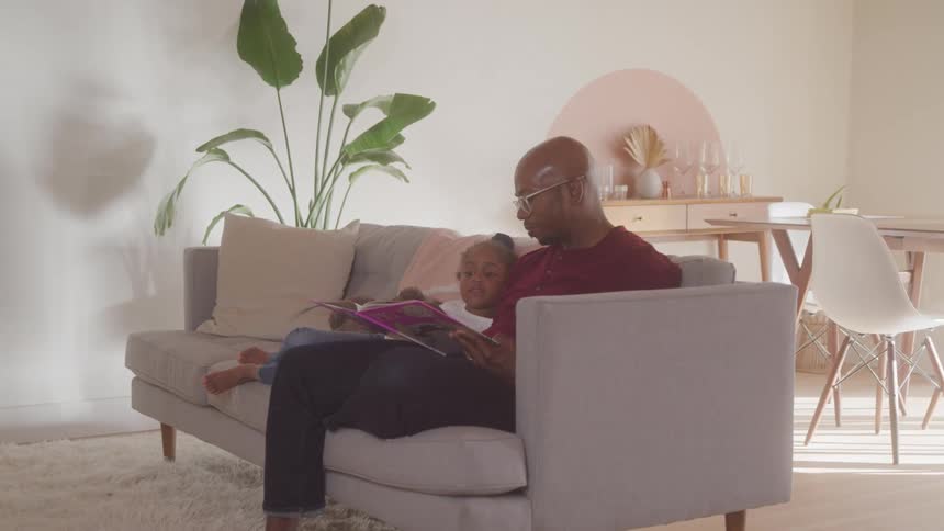 In a brightly lit living room a father wearing glasses and a red shirt reads a book to his young daughter as they sit together on a beige couch