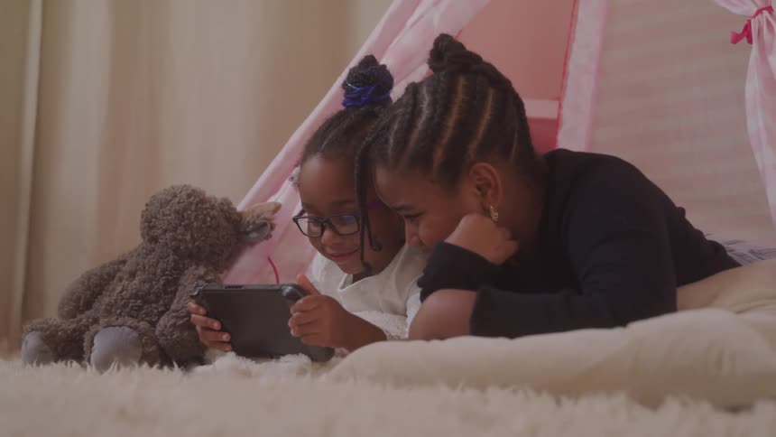Laying sidebyside inside a cozy play tent on a soft rug two young sisters share a moment as one plays a nintendo switch console while the other watches intently