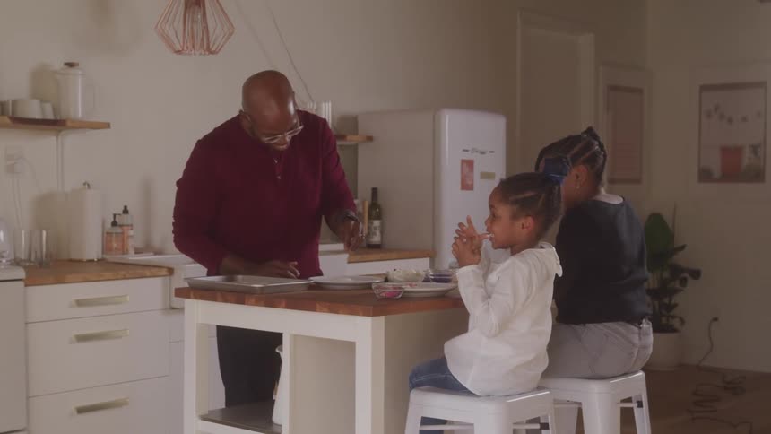 A father serves food to his young daughters while they sit at the kitchen snackbar