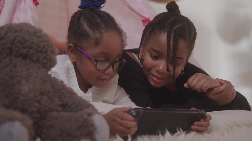 Two young girls are lying on their bedroom floor while looking at an electronic tablet