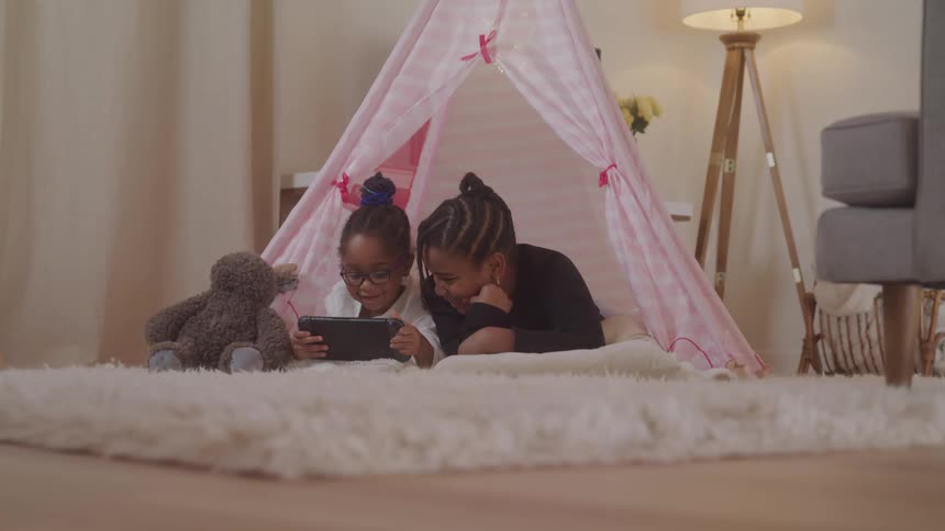 Two children lying in a makeshift tent in the living room interacting with a tablet