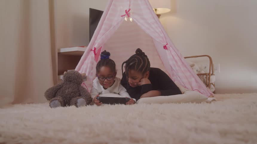 Two children lying on a sheet tent in the living room interacting with a tablet