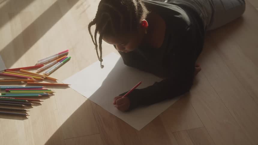 A young person with dark braided hair is lying on a wooden floor illuminated by sunlight drawing on a white piece of paper with a red colored pencil while a collection of various colored pencils and markers is scattered to their left