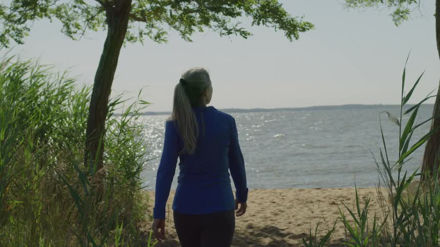 A woman wearing workout clothing walks on a sandy beach on a sunny day