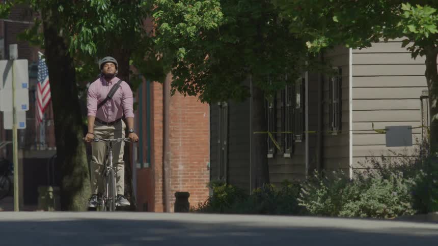 A person wearing a helmet rides a bicycle along a treelined street bordered by buildings and an american flag in the background