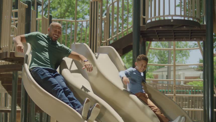 A father in a green shirt and his young son sit sidebyside at the top of parallel playground slides counting down together before they slide down on a sunny day