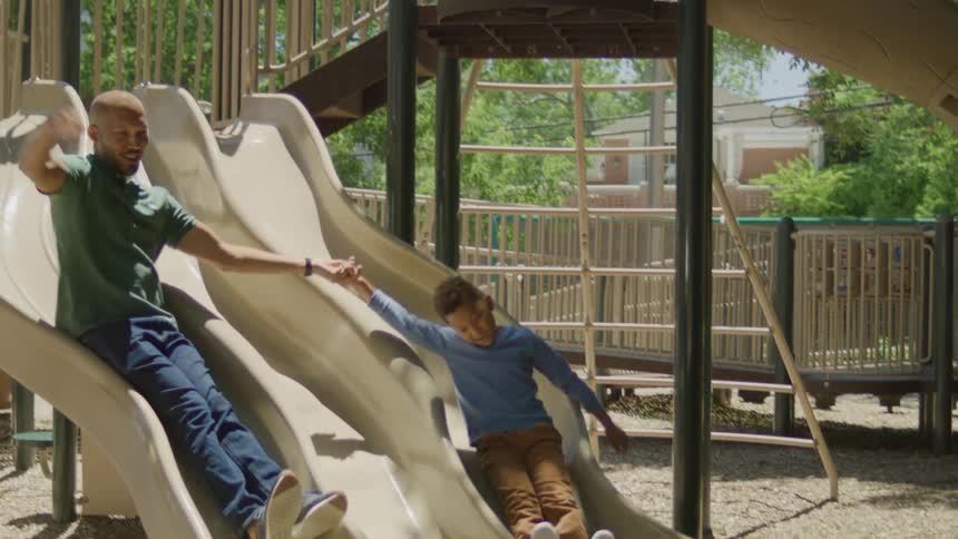 A father and son hold hands while riding down a slide on a playground