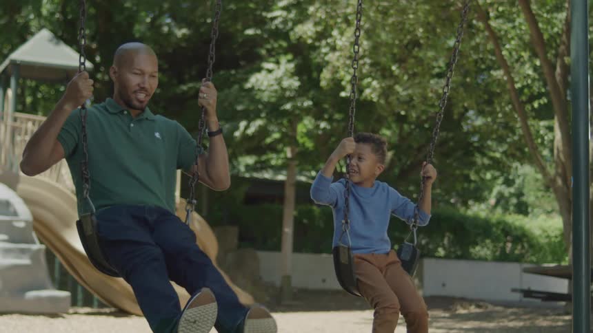 A father and son are joyfully swinging on swings at a playground both beaming with laughter