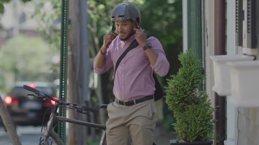 A man wearing a grey helmet a light purple shirt and khaki pants adjusts his collar while standing next to his bicycle on a city street