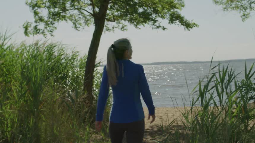 Seen from behind a woman with a blonde ponytail and in a blue sweater and grey leggings walks down a sandy path bordered by tall grass and trees heading towards a large sparkling body of water on a sunny day