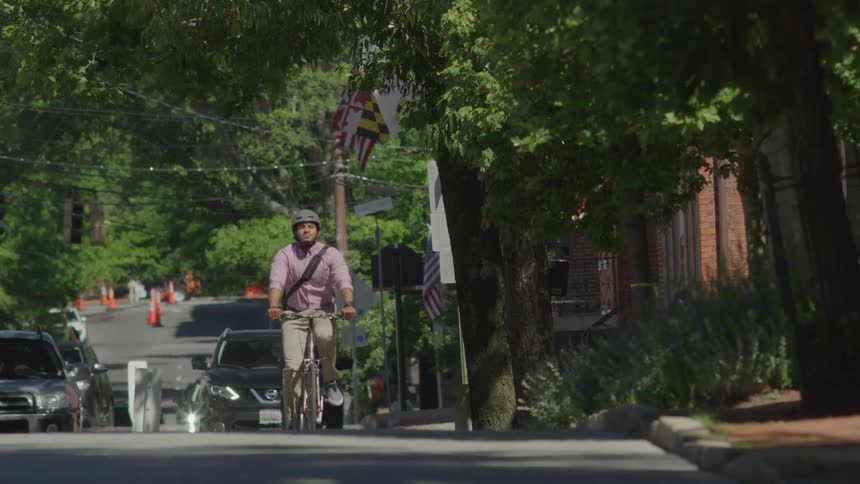 A man wearing a helmet a light pink checkered shirt and light colored pants is riding a bicycle down a tree lined street with parked cars traffic cones and american flags visible