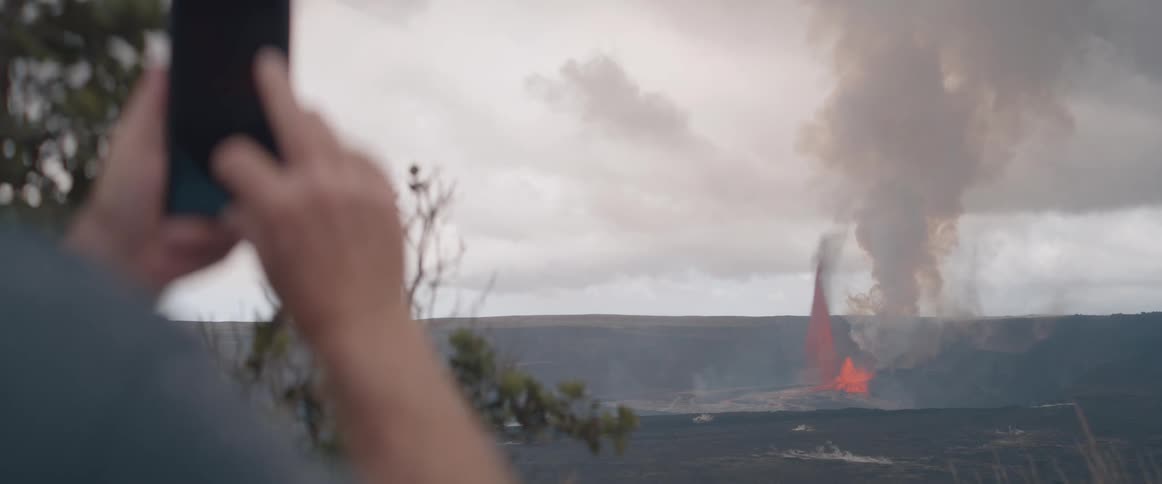Viewed over someones shoulder a smartphone is held up to record a volcanic eruption spewing lava and smoke across a dark landscape