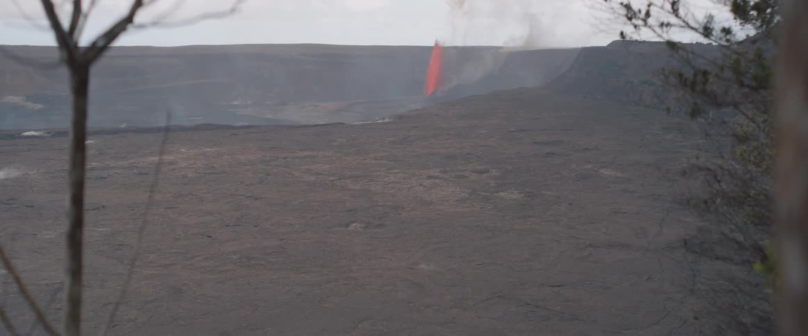 A distant orange lava fountain erupts across a wide barren volcanic landscape under a hazy sky