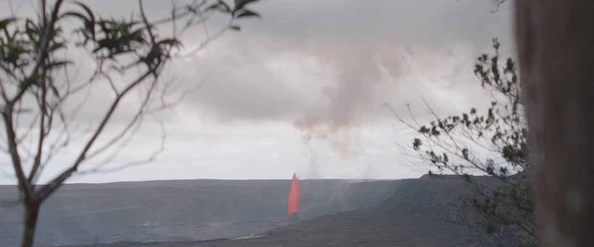 Framed by swaying tree branches in the foreground a distant volcano actively erupts shooting a fountain of bright orange lava and a plume of smoke into the overcast sky