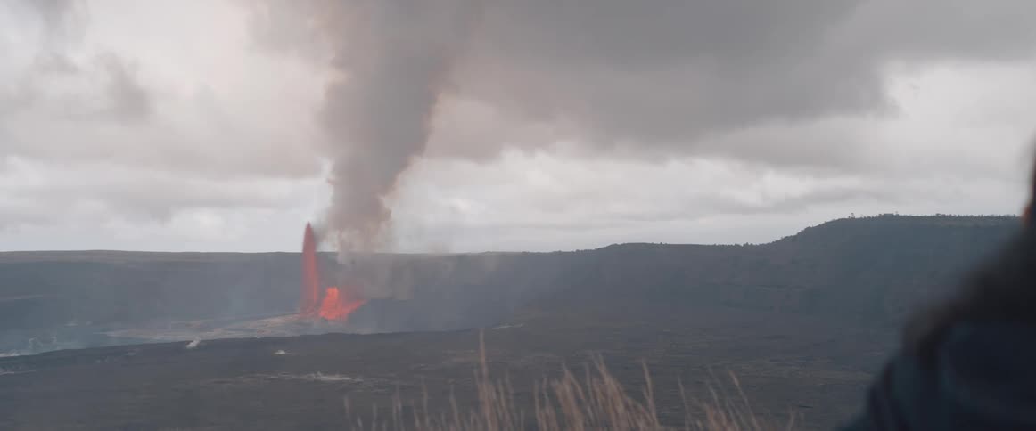Lava fountains and smoke billow from a volcanic eruption across a wide barren landscape under a cloudy sky with dry grass in the foreground
