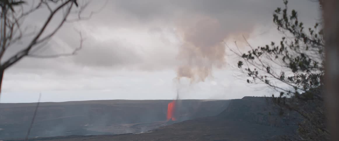 Framed by tree branches swaying gently in the wind a distant volcano actively erupts spewing a fountain of bright orange lava and a plume of smoke into the cloudy sky