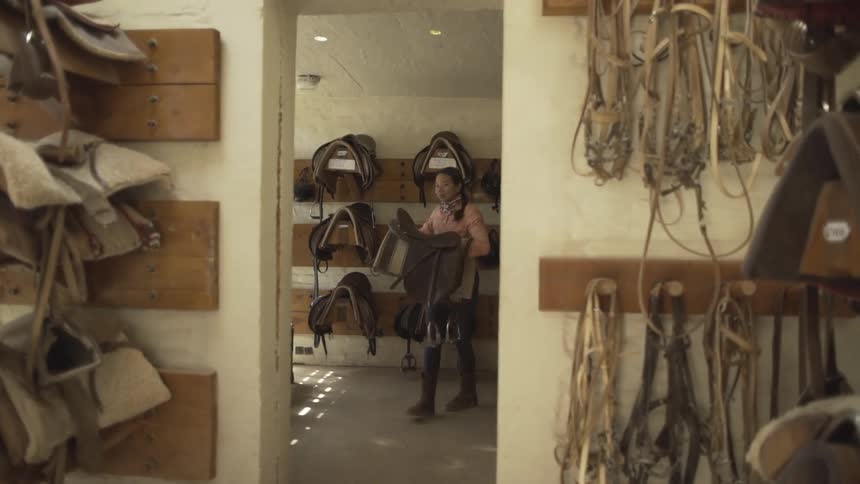 A woman carries a saddle out of the stables