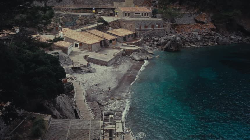 An aerial view captures a quaint coastal village tucked into a rocky cove featuring stone buildings with tile roofs nestled beside clear turquoise water where a person walks