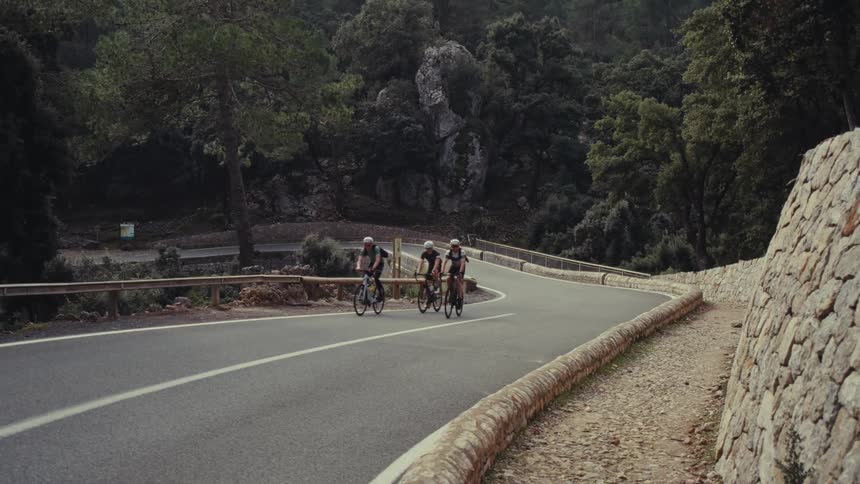 Three cyclists wearing helmets ride their bicycles uphill together around a curve on a paved mountain road bordered by a stone wall and trees