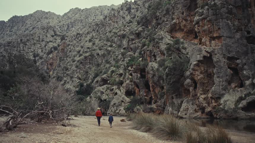 Seen from behind a woman in an orange jacket and a child walk along a dirt path beside a stream at the bottom of a vast rocky canyon