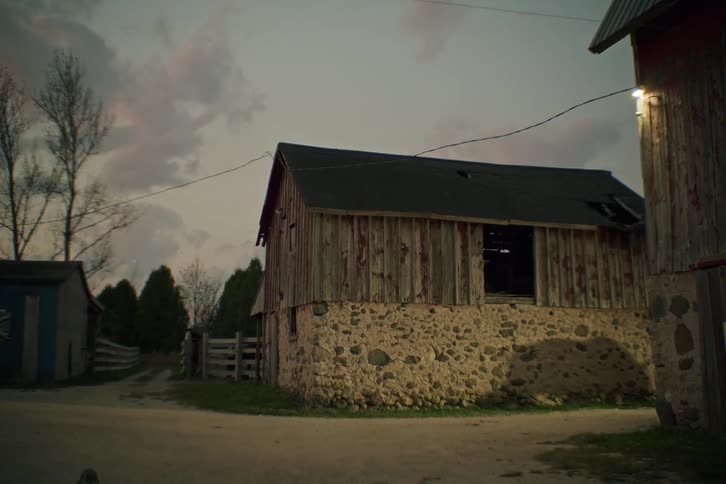 Under a moody cloudy sky at dusk or dawn a person guides a brown horse along a dirt path towards a weathered stone and wood barn on a rural farm property