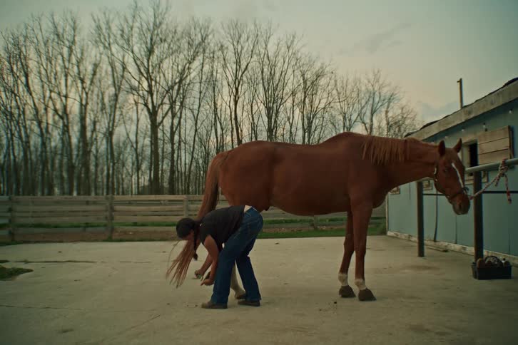 Bending over with her long hair falling forward a woman wearing a black top and jeans lifts and tends to the hind hoof of a brown horse standing on concrete near a stable