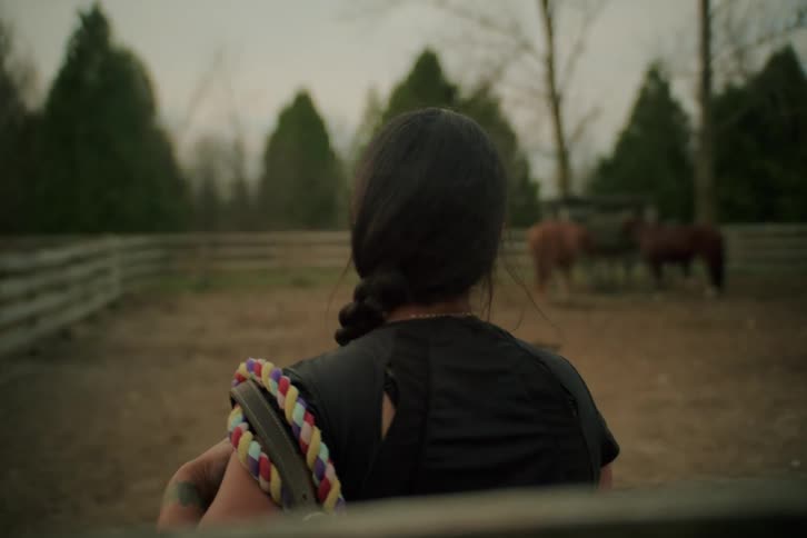 Woman pulls horse tack onto her shoulder while standing in a corral and staring at a small herd of horses during the daytime