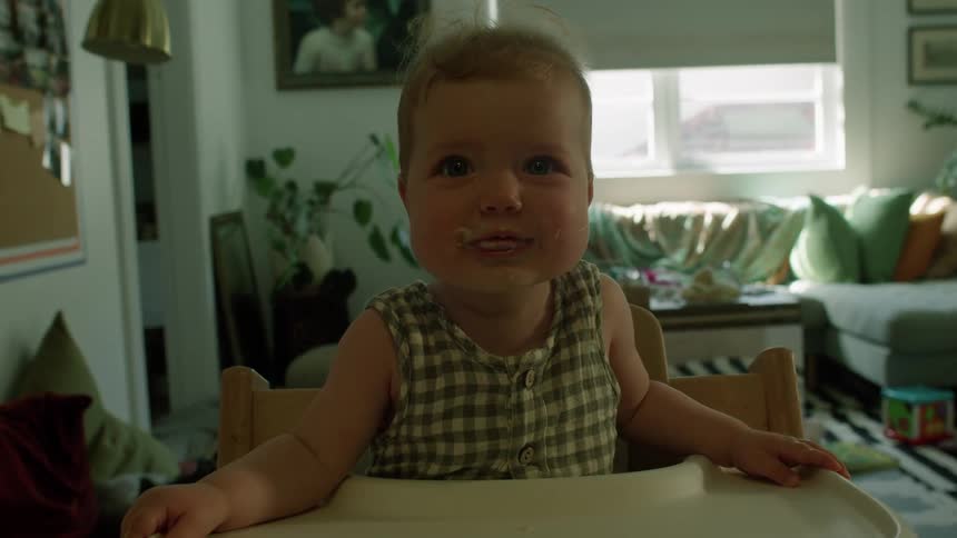 A child smiling with cheeks and lips full of food sits at a wooden table in a cozy living room surrounded by toys