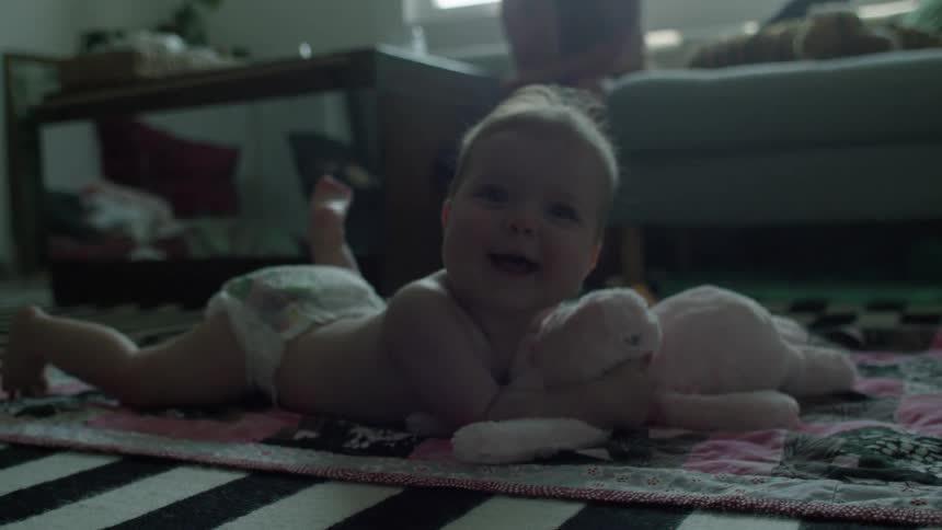 A baby wearing a diaper is lying on their stomach on a patterned rug holding a stuffed animal and looking towards the camera in a dimly lit room