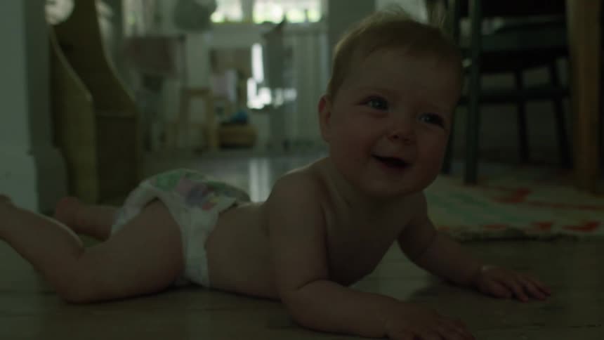 A baby wearing a diaper lies on their stomach on a floor in a softly lit room looking upwards with their mouth open