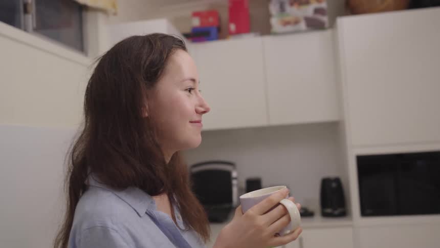 A woman with long brown hair drinks from a white mug in a kitchen setting
