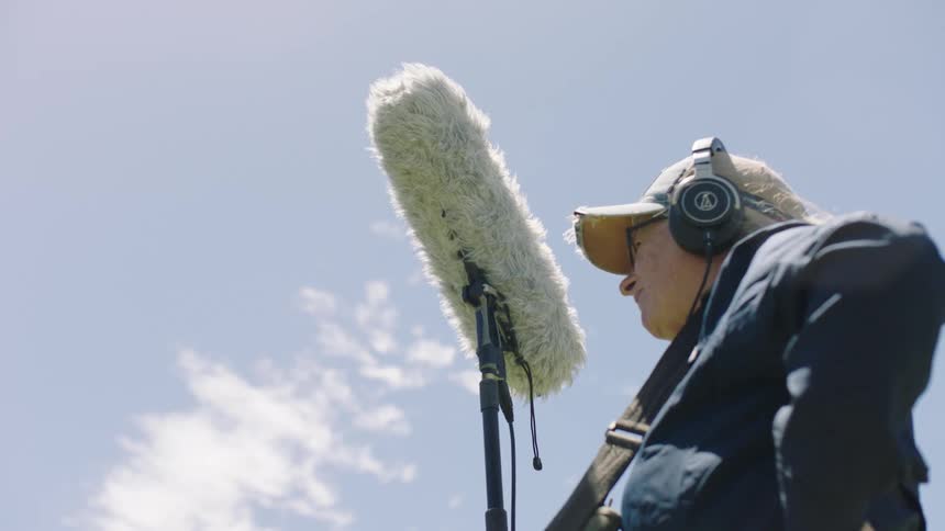 Member of a film crew stands outdoors holding a piece of film equipment on a cloudy day