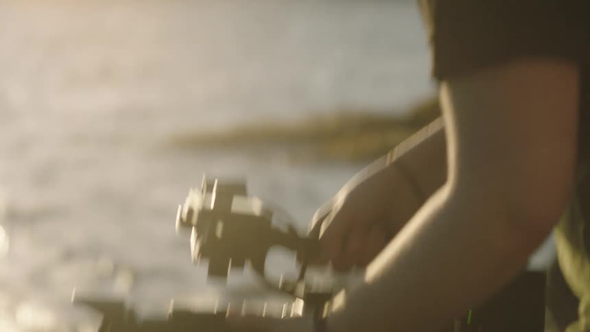 A mans hands are operating a camera stabilizer filming near a body of water with sunlight reflecting off its surface