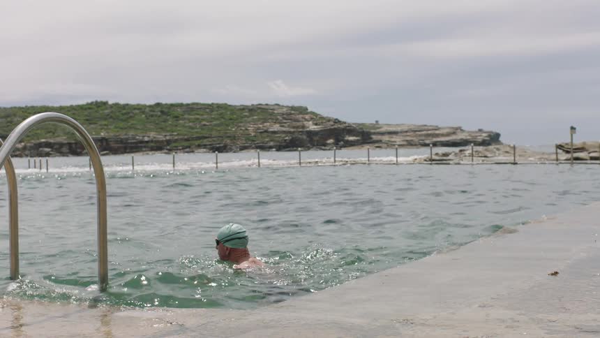 A swimmer is doing the crawl in a large ocean pool enclosed by concrete walls and metal railings with a rocky coastline visible in the background under a cloudy sky