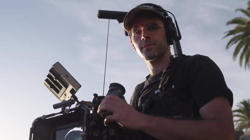 Filmmaker holding a professional video camera in an outdoor setting with palm trees in the background