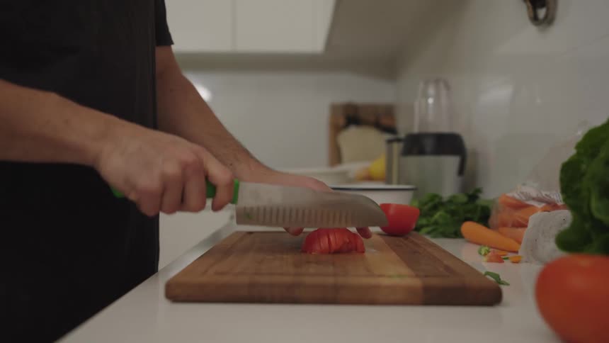 A man wearing a black shirt is in a kitchen using a large knife to chop a red tomato on a wooden cutting board surrounded by other vegetables and kitchen appliances