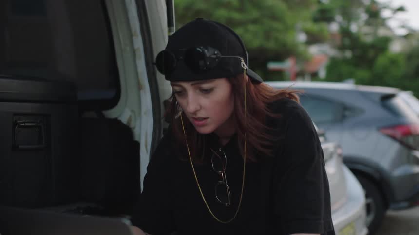 A woman wearing a black cap with goggles perched on top sits at the open back of a van intensely focused on working on a laptop computer