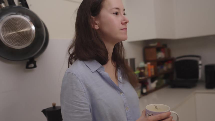 A young woman stands in her kitchen while eating avocado toast and drinking coffee