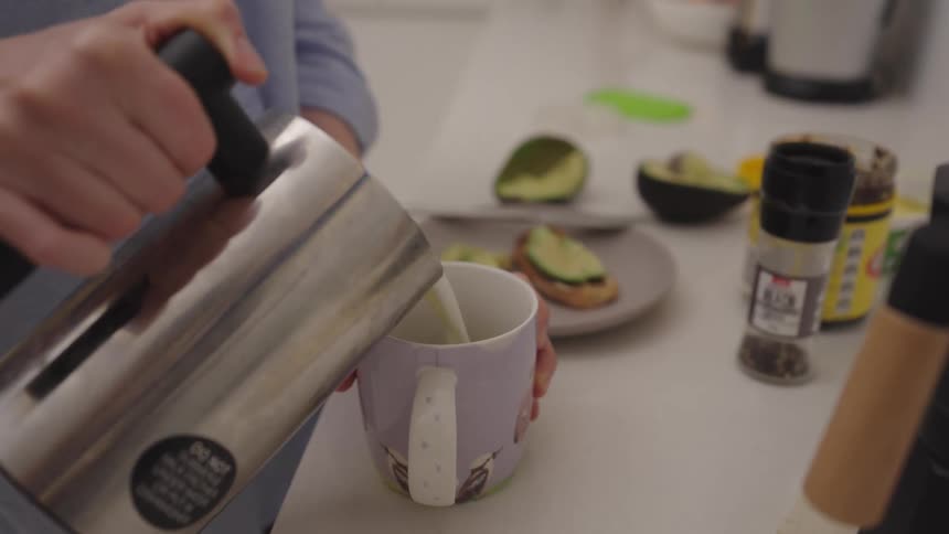 A woman pours coffee from a moka pot adds frothed milk to her mug using a frother and then takes a drink in her kitchen