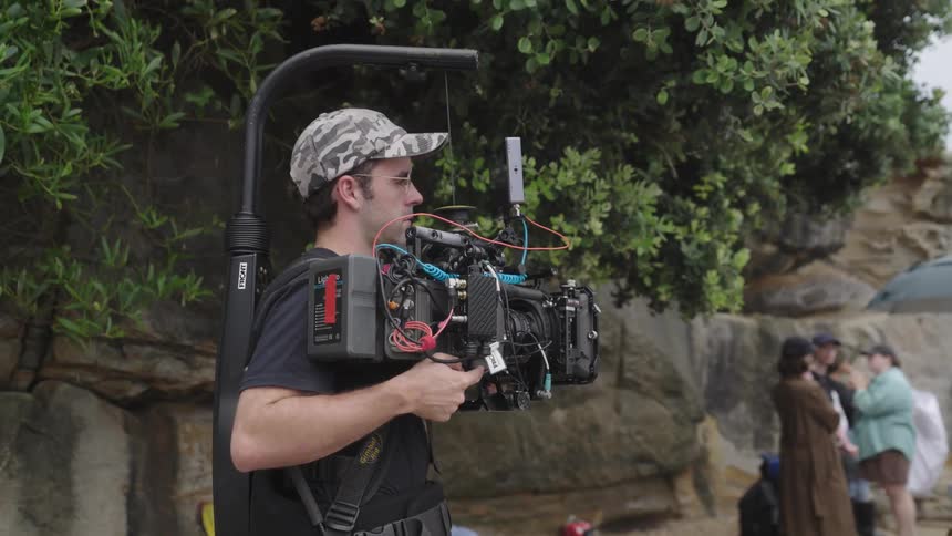 Man using a professional video camera in an outdoor setting with people in the background